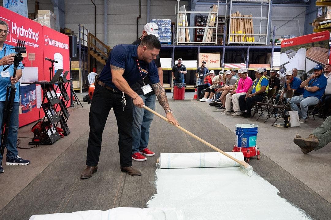 A man rolls a white liquid membrane across the floor with a long handled roller while a seated audience watches.